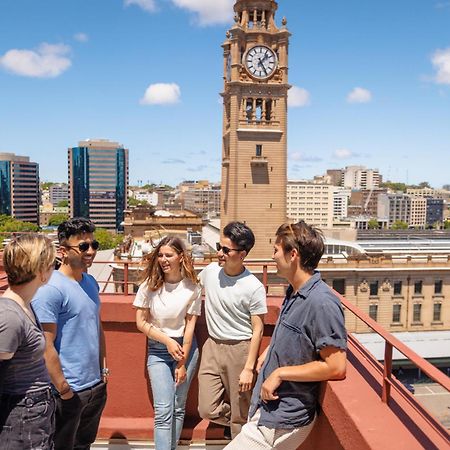 Yha Sydney Central Exterior photo The photo shows a group of five people standing on a rooftop terrace with a view of a cityscape. In the background, there is a clock tower and several modern buildings. The sky is clear with a few clouds. The people are casually dressed and appear to