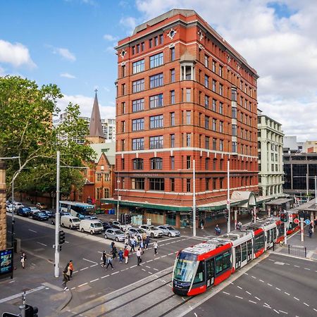 Yha Sydney Central Exterior photo The photo shows a bustling urban scene, featuring a prominent building with a red brick facade. The structure stands several stories tall and is located at a street intersection. In the foreground, a tram is seen on the tracks, moving along the stree