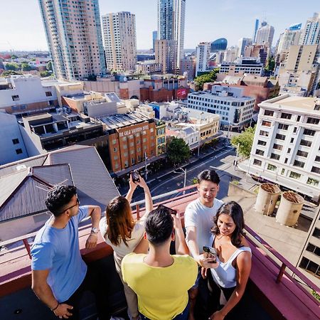 Yha Sydney Central Exterior photo The photo shows a group of five people on a rooftop, enjoying a view of a city skyline. They are standing close together, engaged in conversation or taking photos. The background features various tall buildings and a bustling urban area, indicating a