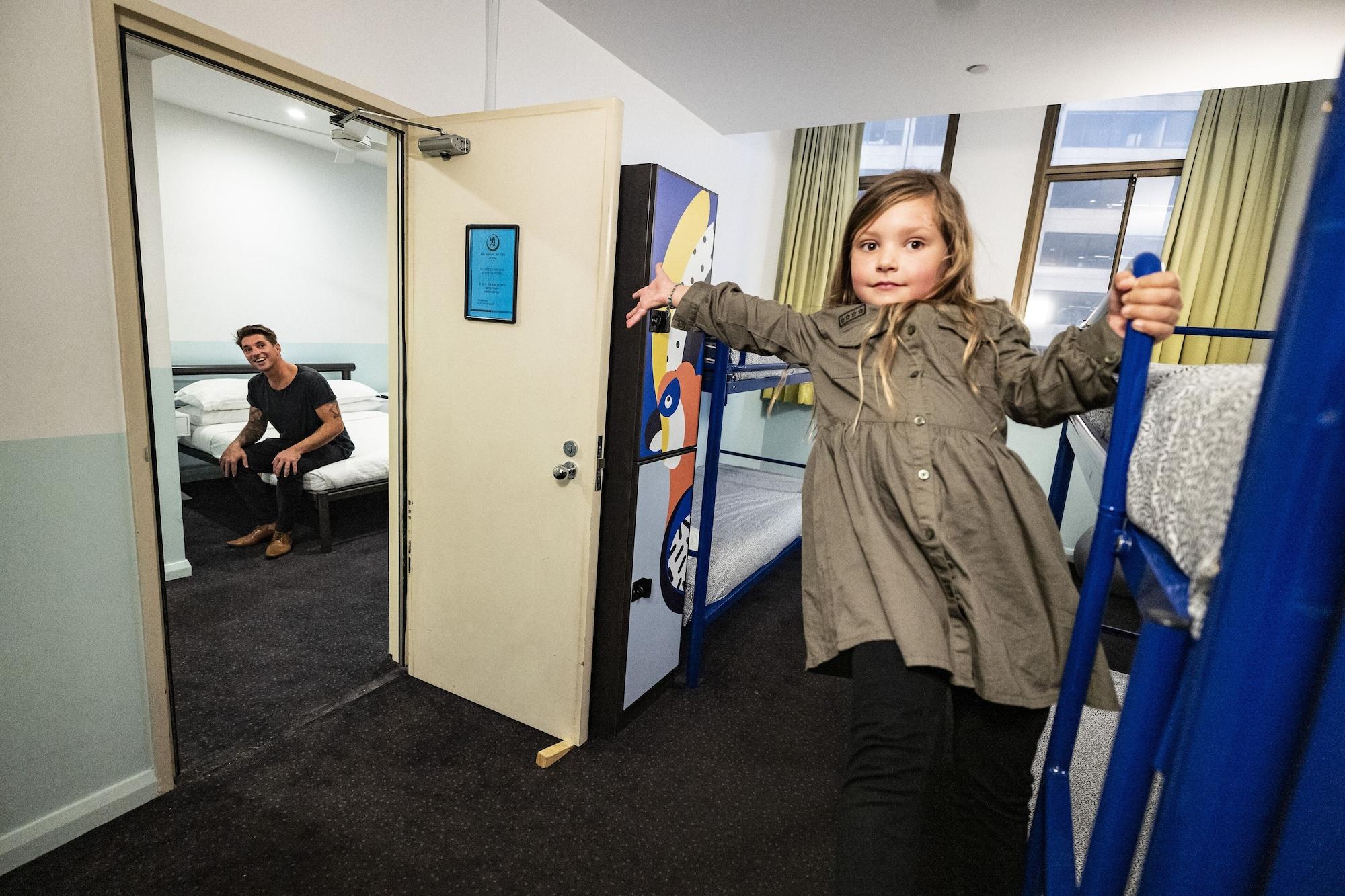 Yha Sydney Central Exterior photo The photo shows a child wearing a light grey coat, playfully posing as she leans against a blue bunk bed in a shared space. In the background, there is an open door leading to another room where a young man is seated on a bed, looking towards the cam