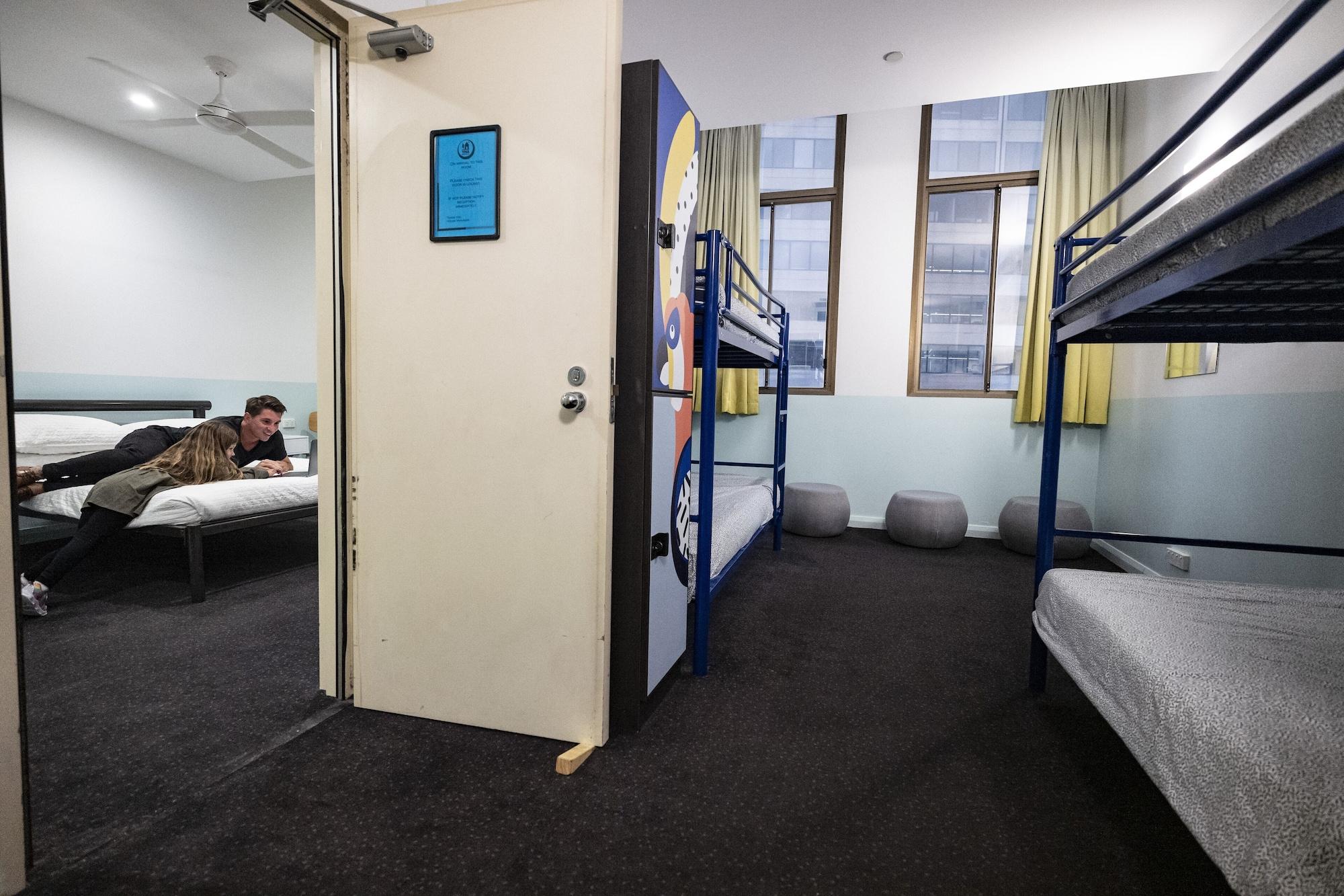 Yha Sydney Central Exterior photo The photo depicts a room that appears to be a hostel or dormitory. On one side, there is a single bed with a person sitting on it, using a device, possibly a laptop or phone. Adjacent to this, there is an open door leading to another space. On the op