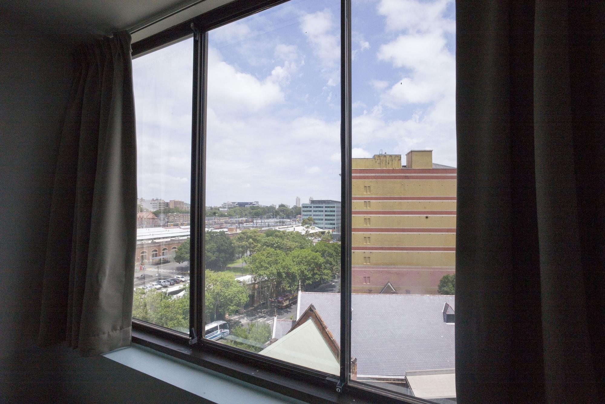Yha Sydney Central Exterior photo The photo shows a view from a window featuring a large glass window with multiple panes. Outside, there is a cityscape with buildings, including a large yellow structure. The sky appears bright with a few clouds, indicating a sunny day. Trees and gre