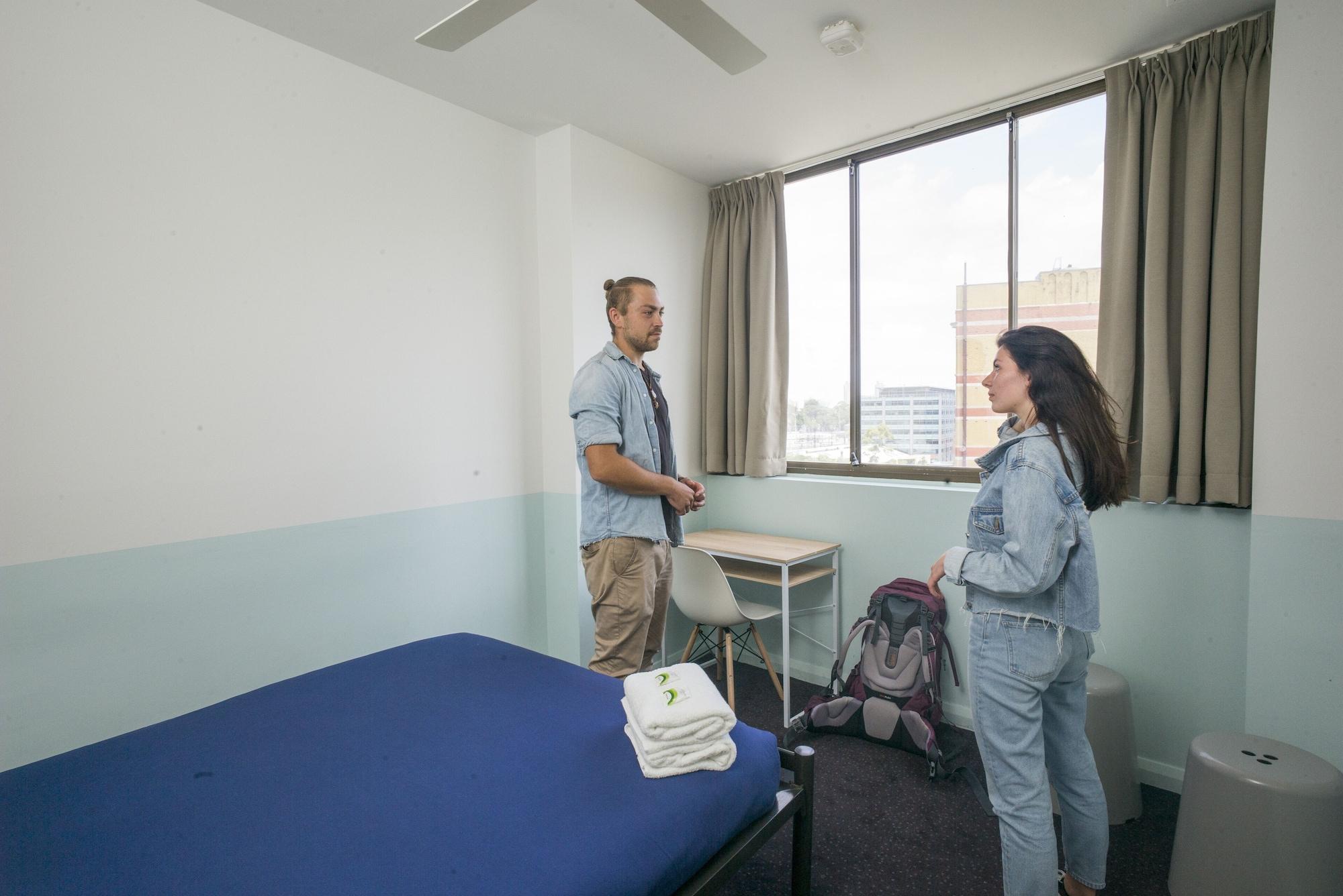 Yha Sydney Central Exterior photo The photo shows a simple, modern room, likely in a hostel or shared accommodation. It features a bed with blue bedding and neatly stacked white towels. There is a window with curtains, offering a view of the outside. Two people are standing in the ro