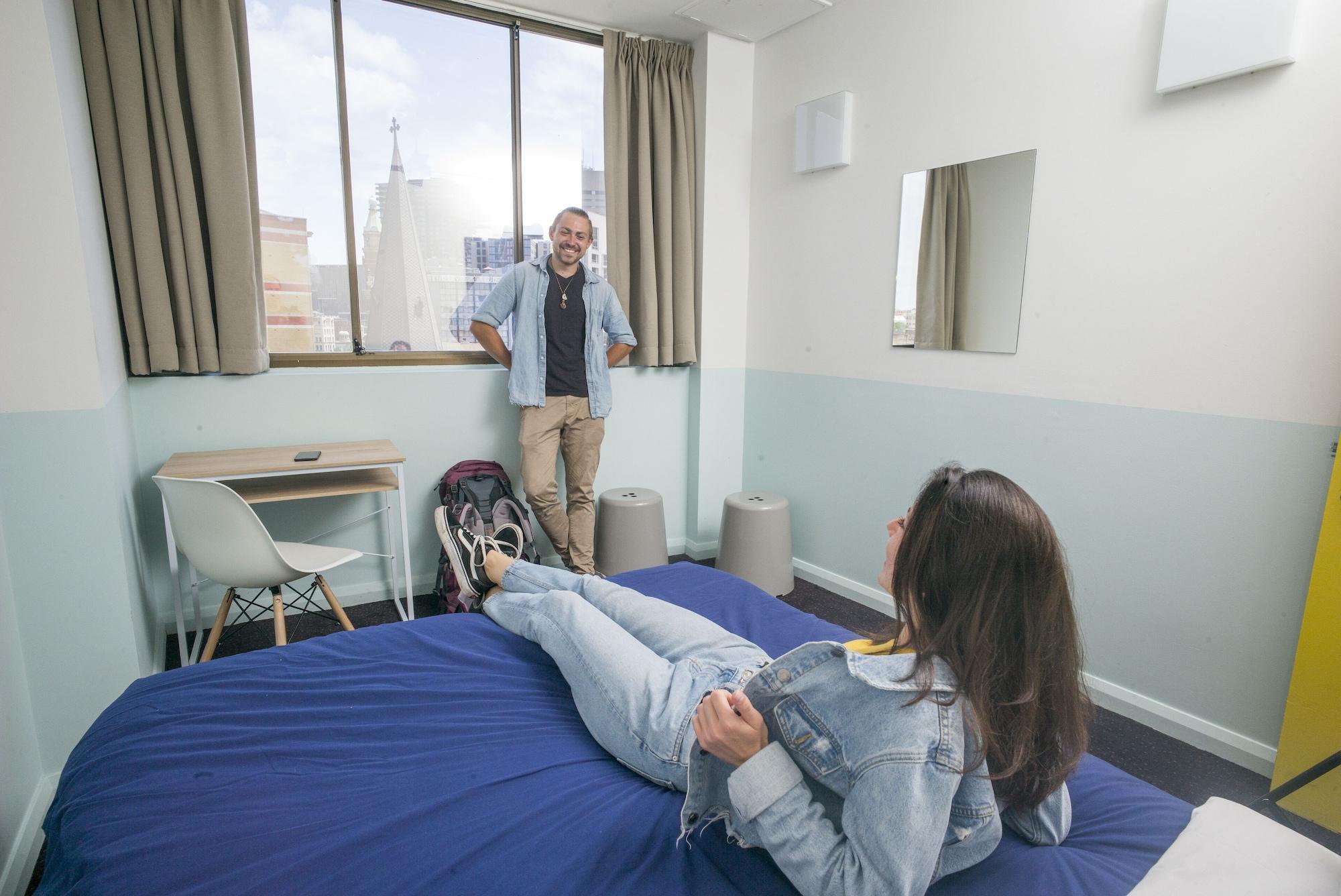 Yha Sydney Central Exterior photo The photo shows a room with a modern, minimalistic design. A man is standing near a window, smiling, while a woman is reclining on a bed, looking towards him. The bed has a blue cover, and there’s a small table and chair visible in the room. The wall