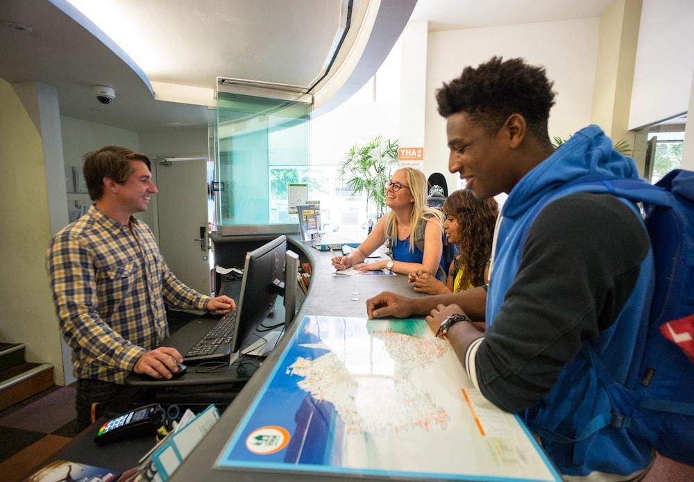 Yha Sydney Central Exterior photo The photo depicts a scene at a reception desk, likely in a hotel or travel agency. There is a male staff member, smiling, who is interacting with a group of three individuals. The staff member is standing behind a counter with a computer and appears 