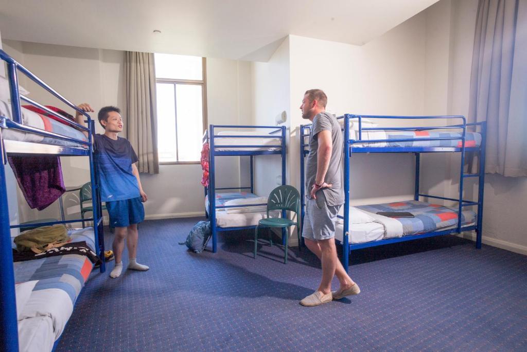 Yha Sydney Central Exterior photo The photo shows a room with two individuals engaged in conversation. The room features several blue metal bunk beds, arranged in a way that suggests a dormitory or shared living space. There are two empty bunks, and a green chair is positioned betwee