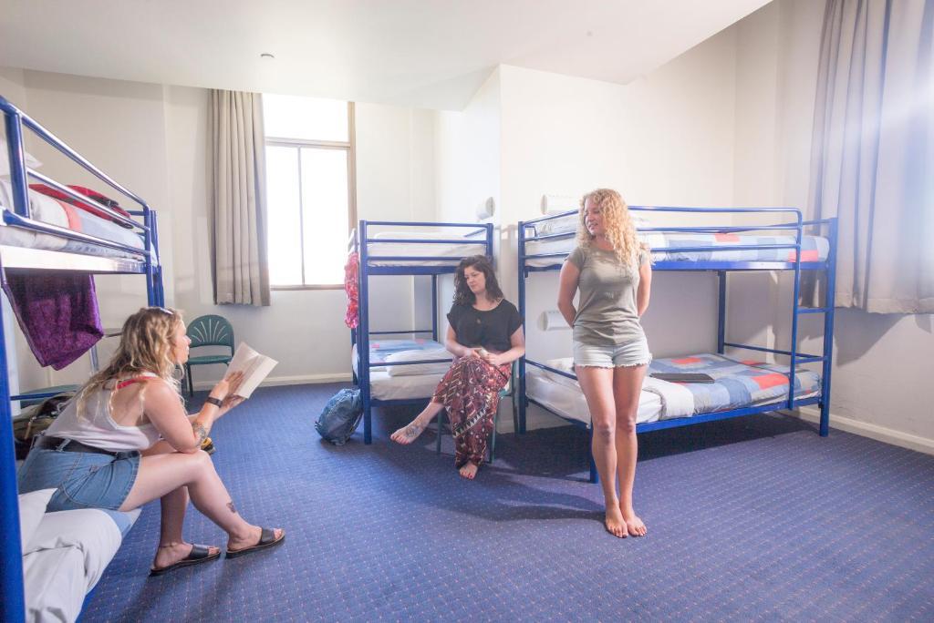 Yha Sydney Central Exterior photo The photo shows a hostel room with three women and bunk beds. Two of the women are seated; one is reading a book while the other is sitting and looking at the camera. The third woman is standing, facing the camera. The room has blue carpeting and lar