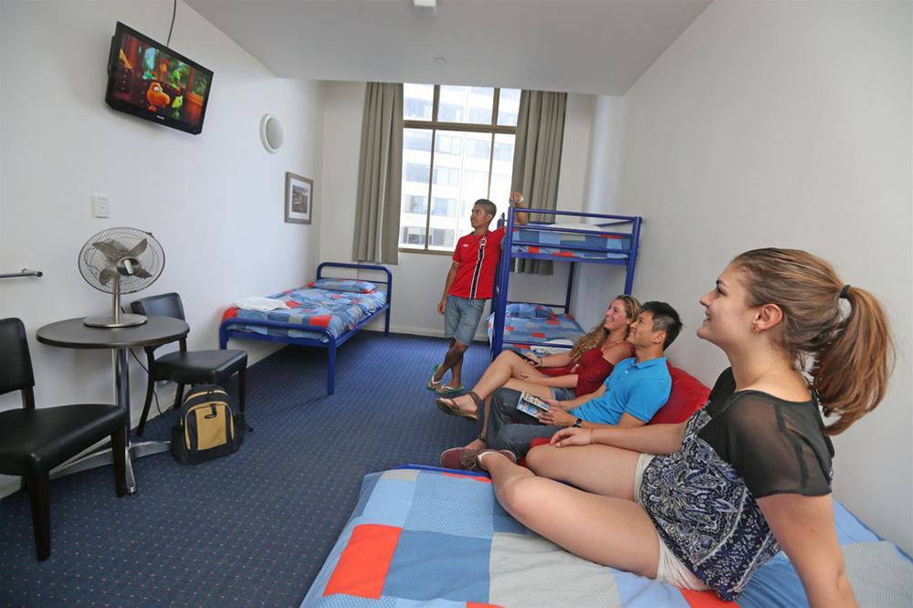 Yha Sydney Central Exterior photo The photo shows a group of four young people enjoying a moment together in a room. They are sitting on a bed and watching a TV mounted on the wall, which displays a colorful image. The group includes two males and two females, with one male standing 