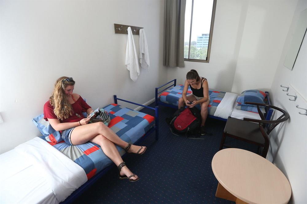 Yha Sydney Central Exterior photo The photo shows a simple hotel room with two single beds covered in colorful blankets. There are two women sitting in the room; one is sitting on the edge of one bed, looking at her phone, while the other is seated on the floor, possibly reading or u
