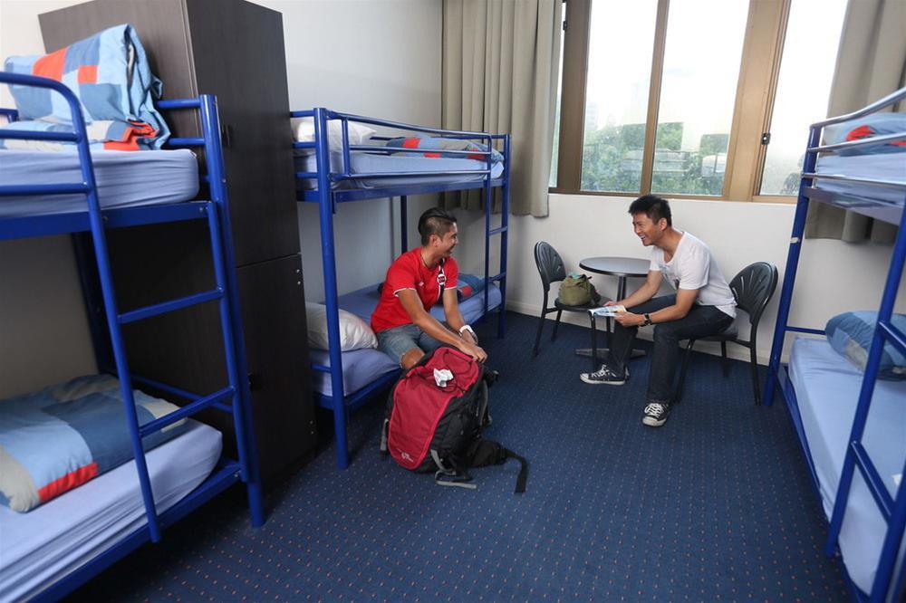 Yha Sydney Central Exterior photo The photo shows a dormitory room with two bunk beds arranged along the walls. The beds have blue frames and are made up with light blue bedding. In the room, two young men are seated: one is on a chair at a small table, while the other is sitting on 
