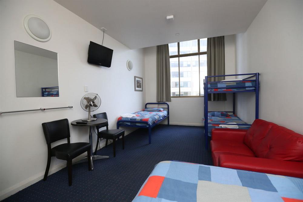 Yha Sydney Central Exterior photo The photo shows a small hotel or hostel room. Features include:

- A window with natural light coming in, partially covered by curtains.
- A flat-screen TV mounted on the wall.
- Two sets of bunk beds, indicating capacity for multiple guests.
- A sma