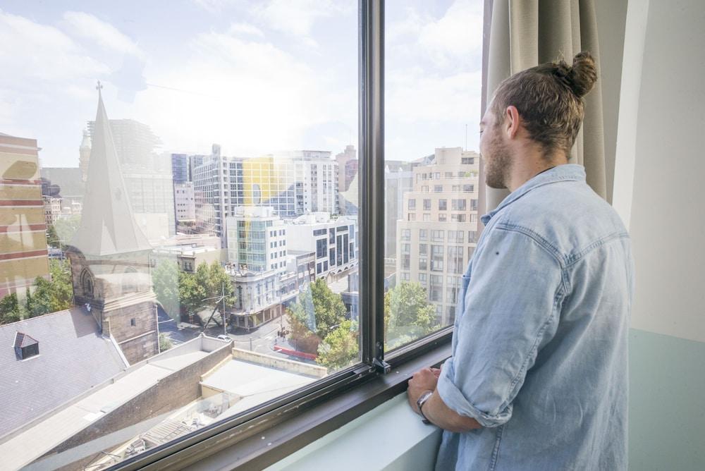 Yha Sydney Central Exterior photo The photo shows a person standing by a large window and looking out at a cityscape. The individual appears to have long hair tied in a bun and is wearing a light-colored, casual shirt. Outside the window, there are various buildings, including a mix 