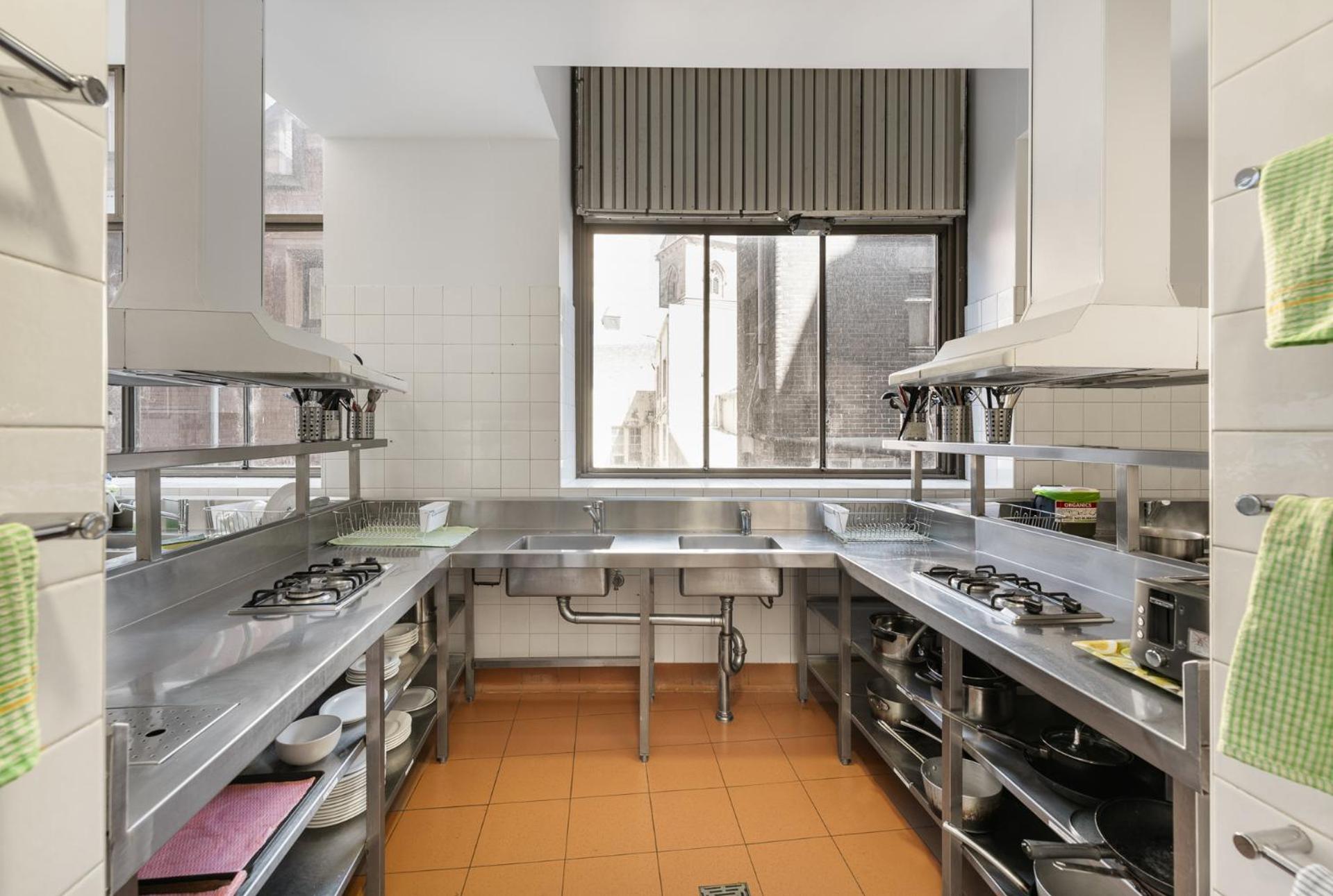 Yha Sydney Central Exterior photo The photo shows a modern kitchen space with stainless steel countertops and appliances. There are multiple cooking stations, each equipped with gas burners and exhaust hoods. The floor is tiled in a warm orange color, contrasting with the silver of t