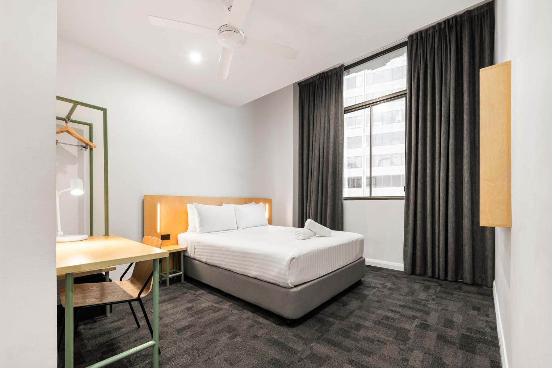 Yha Sydney Central Exterior photo The photo shows a modern hotel room with a cozy and minimalistic design. There is a double bed with white linens, a bedside lamp, and a small decorative pillow. To the side, there is a simple wooden desk and a chair. A standing clothing rack is visib