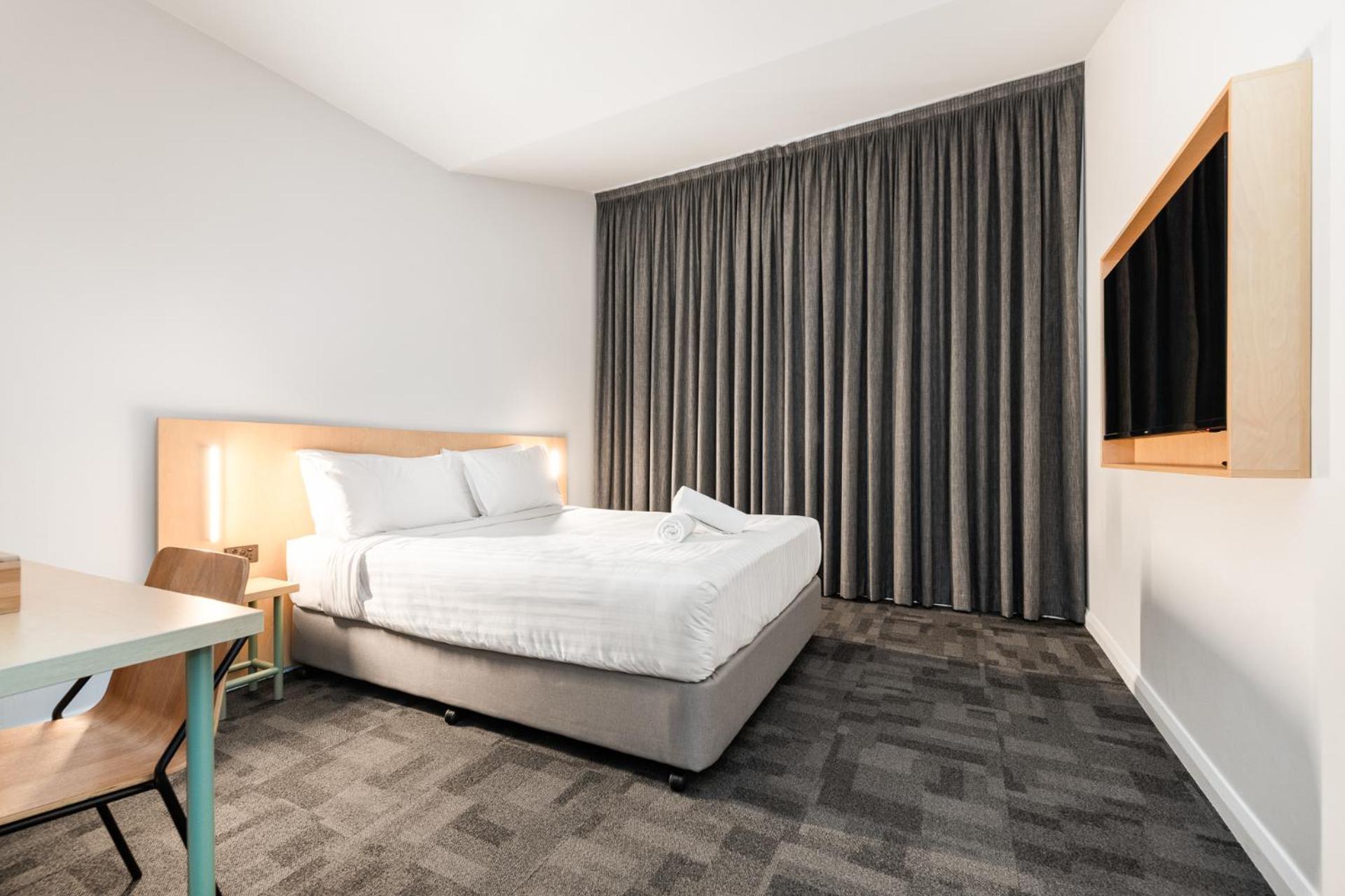 Yha Sydney Central Exterior photo The photo shows a modern hotel room. There is a bed with white bedding positioned against a light-colored wall. Next to the bed, there's a small bedside table with a lamp. A simple desk and chair are also present in the room. The large window is cove