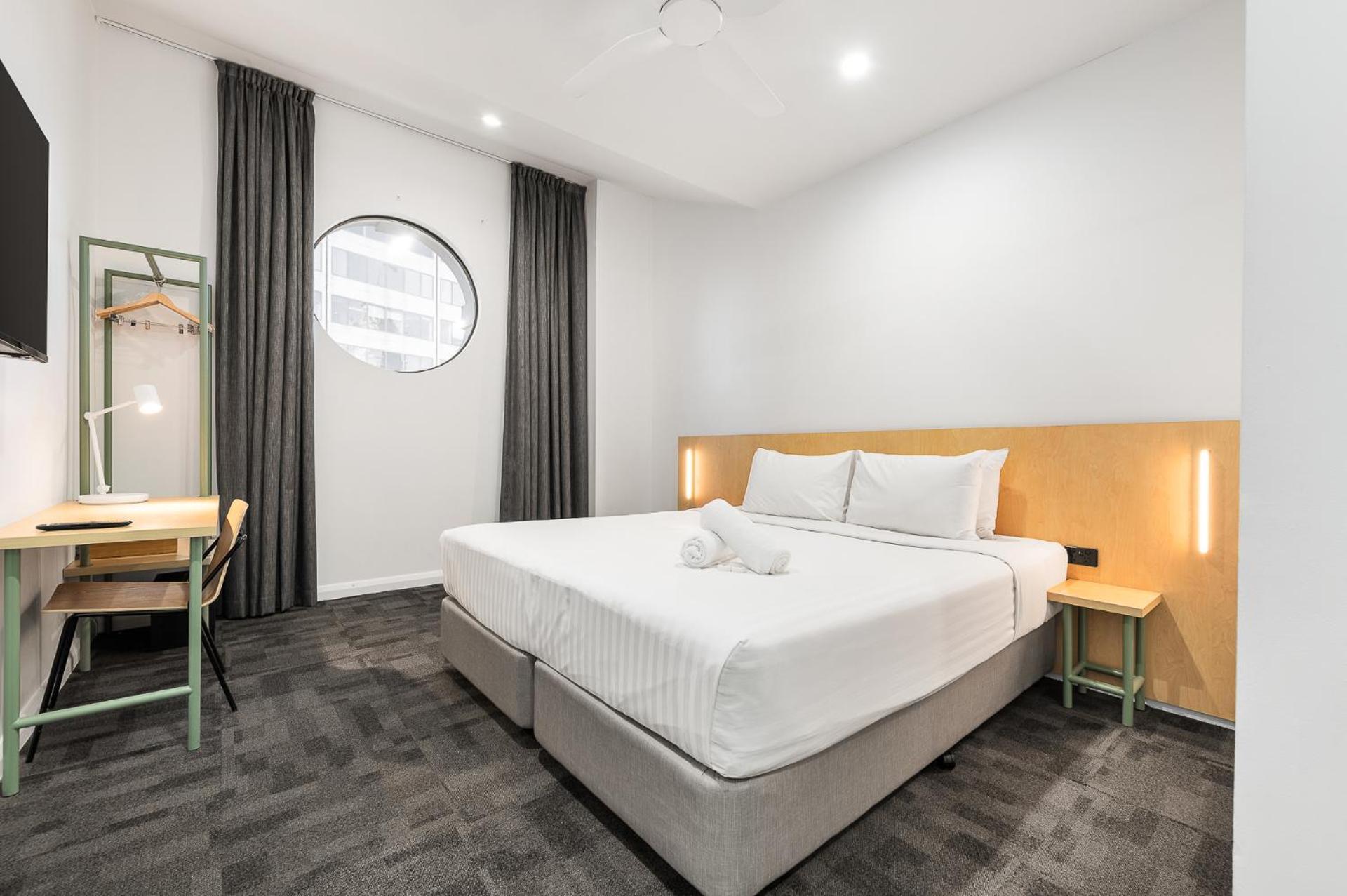 Yha Sydney Central Exterior photo The photo shows a modern hotel room featuring a large bed with white bedding, accompanied by two pillows. To one side of the bed, there is a small nightstand with a lamp. The room has a round window that lets in natural light, framed by floor-length 