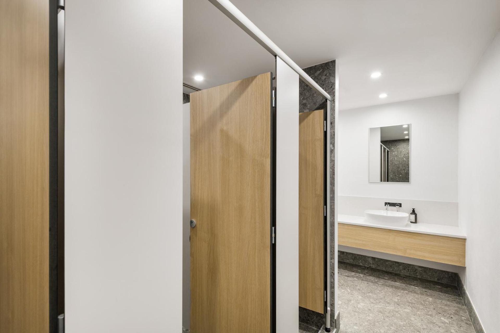 Yha Sydney Central Exterior photo The photo shows a modern restroom with several features. To the left, there are partial partitions for individual stalls, featuring wooden doors that provide privacy. The stalls are separated by white panels. On the right side, there is a sink area w