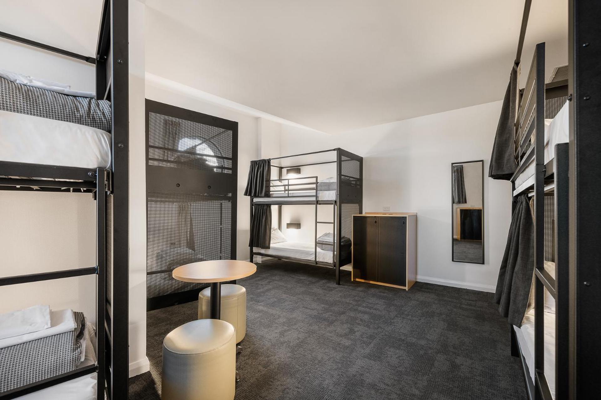 Yha Sydney Central Exterior photo The photo shows a modern shared dormitory room. It features several metal bunk beds with privacy curtains. The floor is carpeted in dark gray, and there are minimalistic wooden furniture pieces, including a small round table and two stools. A tall ca
