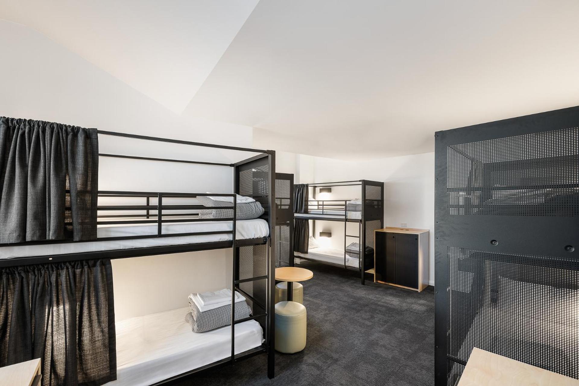 Yha Sydney Central Exterior photo The image shows a modern dormitory-style room featuring bunk beds. There are two sets of black metal bunk beds, each with a ladder for access to the upper bunks. The beds have white bedding, and curtains are drawn for privacy. In the center of the ro