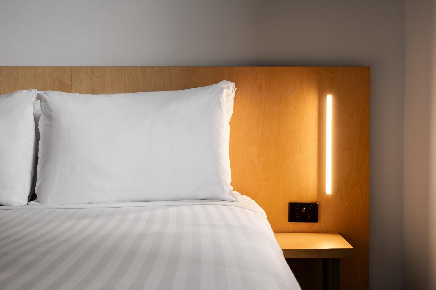 Yha Sydney Central Exterior photo The photo shows a neatly made bed with a white pillow and a white bedding set. The headboard is wooden with a smooth finish, and there is a narrow, modern light fixture mounted on the wall next to the bed. Below the light, there is a small nightstand