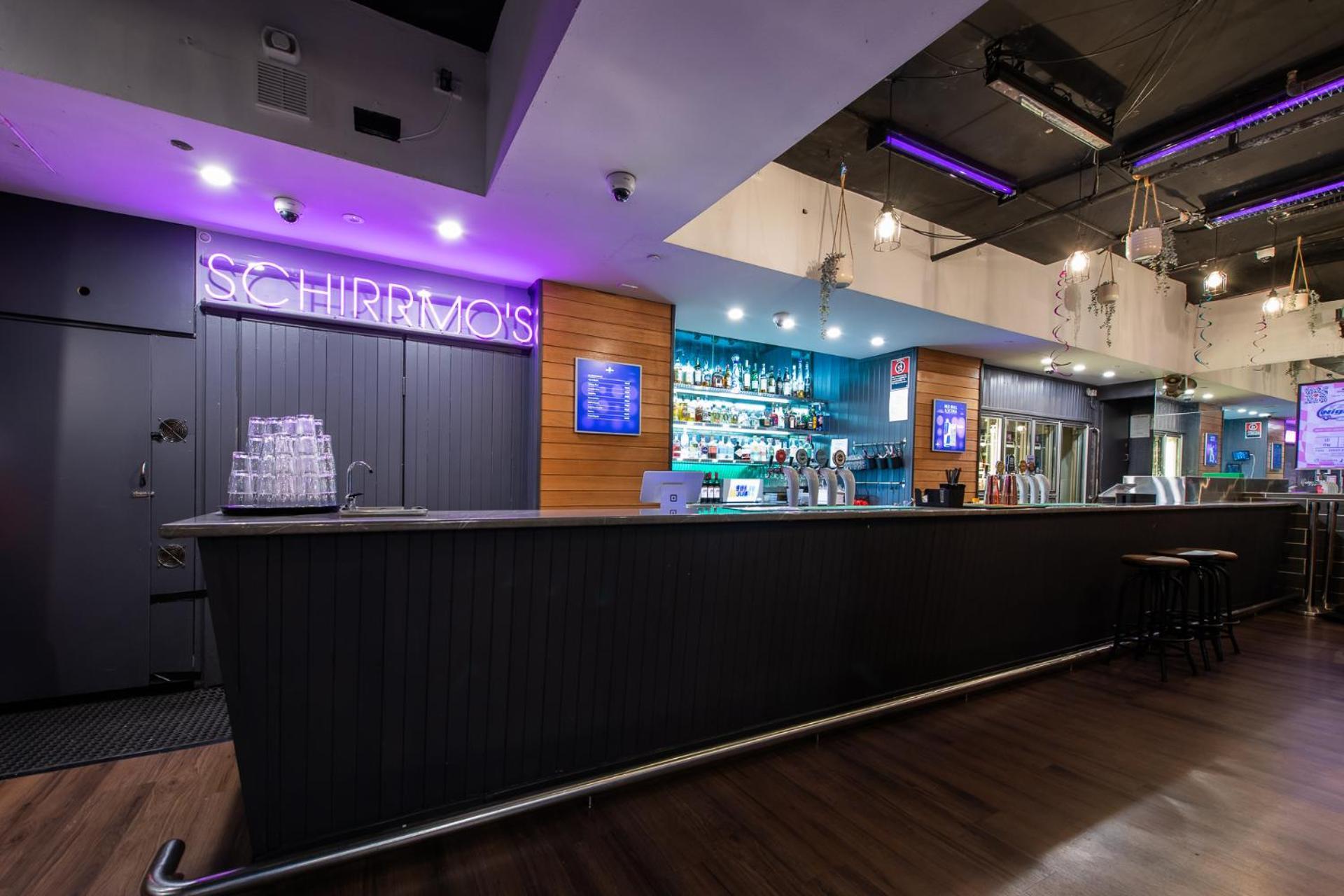 Yha Sydney Central Exterior photo The photo shows a modern bar interior. The bar is long and sleek with a dark countertop. Behind the bar, there are shelves displaying various bottles and drinks, illuminated by blue lighting. A neon sign reading "SCHIRRVOIS" adds a vibrant touch to t