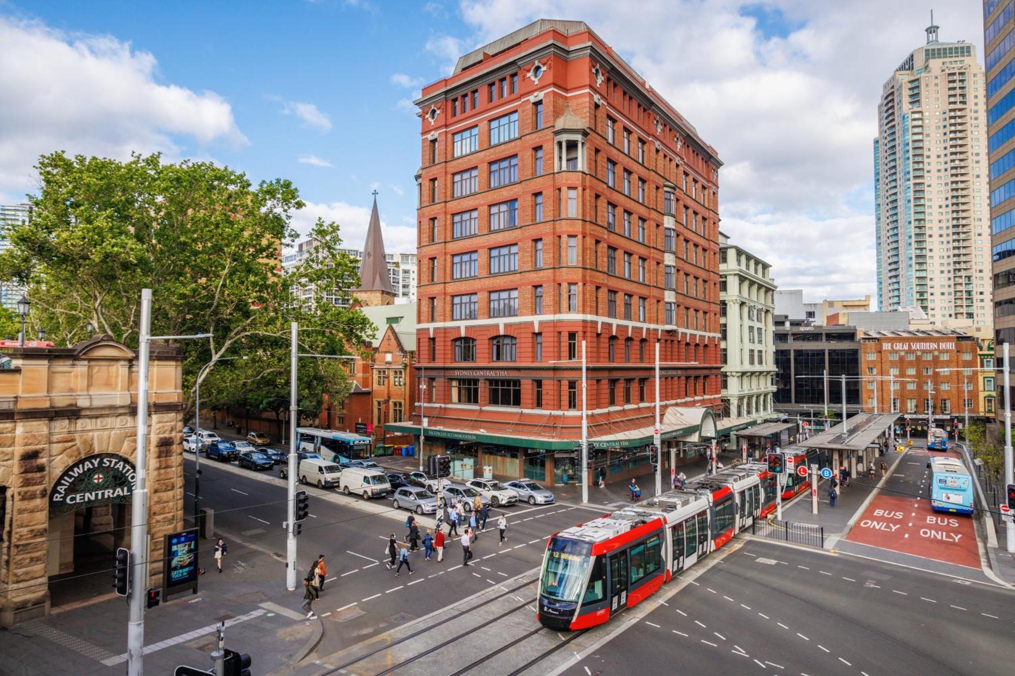 Yha Sydney Central Exterior photo The photo shows a bustling urban scene, featuring a prominent building with a red brick facade. The structure stands several stories tall and is located at a street intersection. In the foreground, a tram is seen on the tracks, moving along the stree