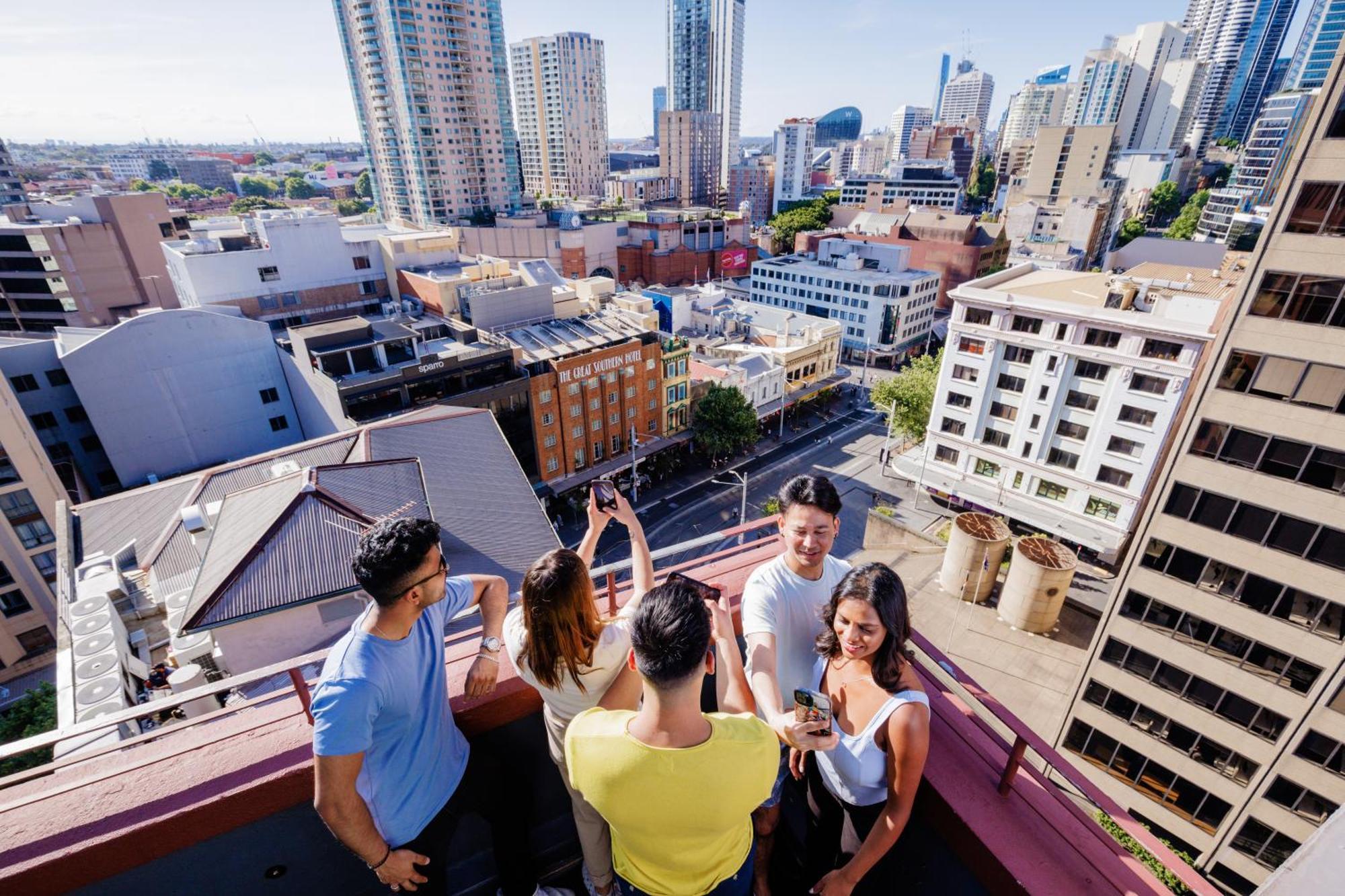 Yha Sydney Central Exterior photo The photo shows a group of five people on a rooftop, enjoying a view of a city skyline. They are standing close together, engaged in conversation or taking photos. The background features various tall buildings and a bustling urban area, indicating a