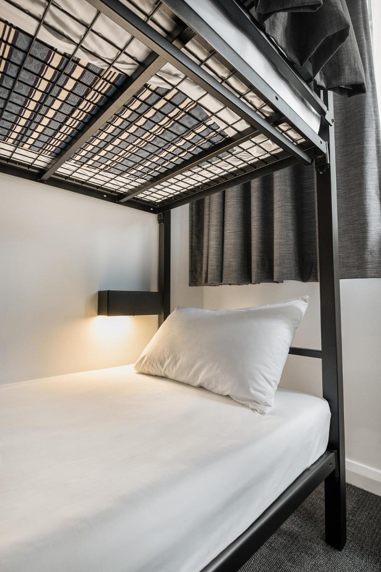 Yha Sydney Central Exterior photo The image shows a close-up view of a bunk bed. The lower bunk features a white mattress with a neatly arranged white pillow. A small light fixture is attached to the side of the bed, providing illumination. In the background, there are dark curtains 