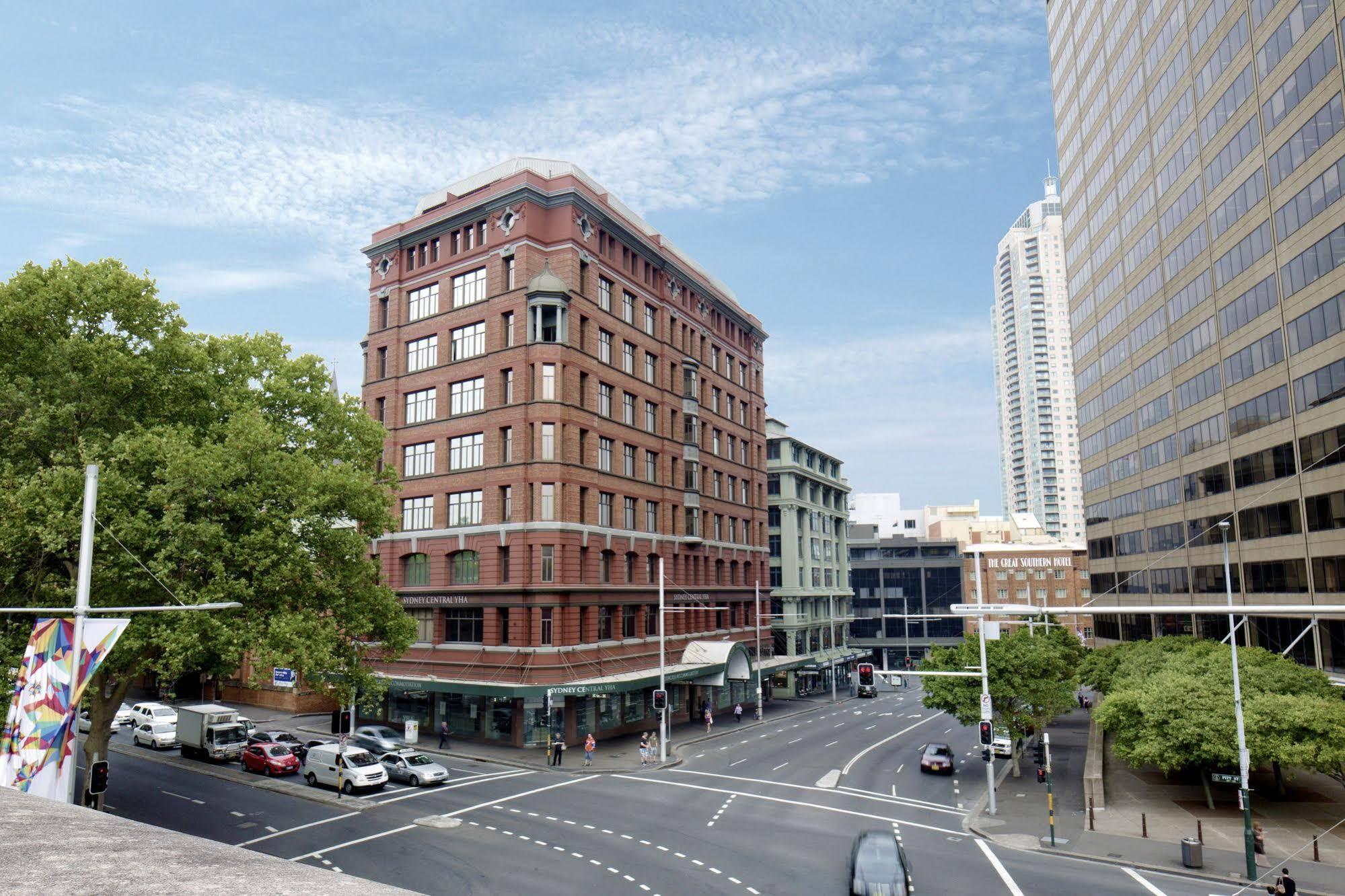 Yha Sydney Central Exterior photo The photo depicts a cityscape featuring a prominent brick building with several stories, characterized by its historical architectural style. It's situated at the intersection of two streets, with traffic visible in the foreground. On the left, there