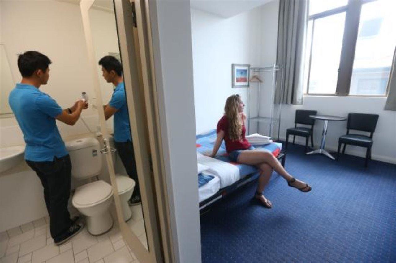 Yha Sydney Central Exterior photo The photo depicts a small room that appears to be a modest hotel or hostel accommodation. On one side, there's a person standing by a bathroom area, visible with a toilet and sink, seemingly preparing to do something with a water bottle. On the other