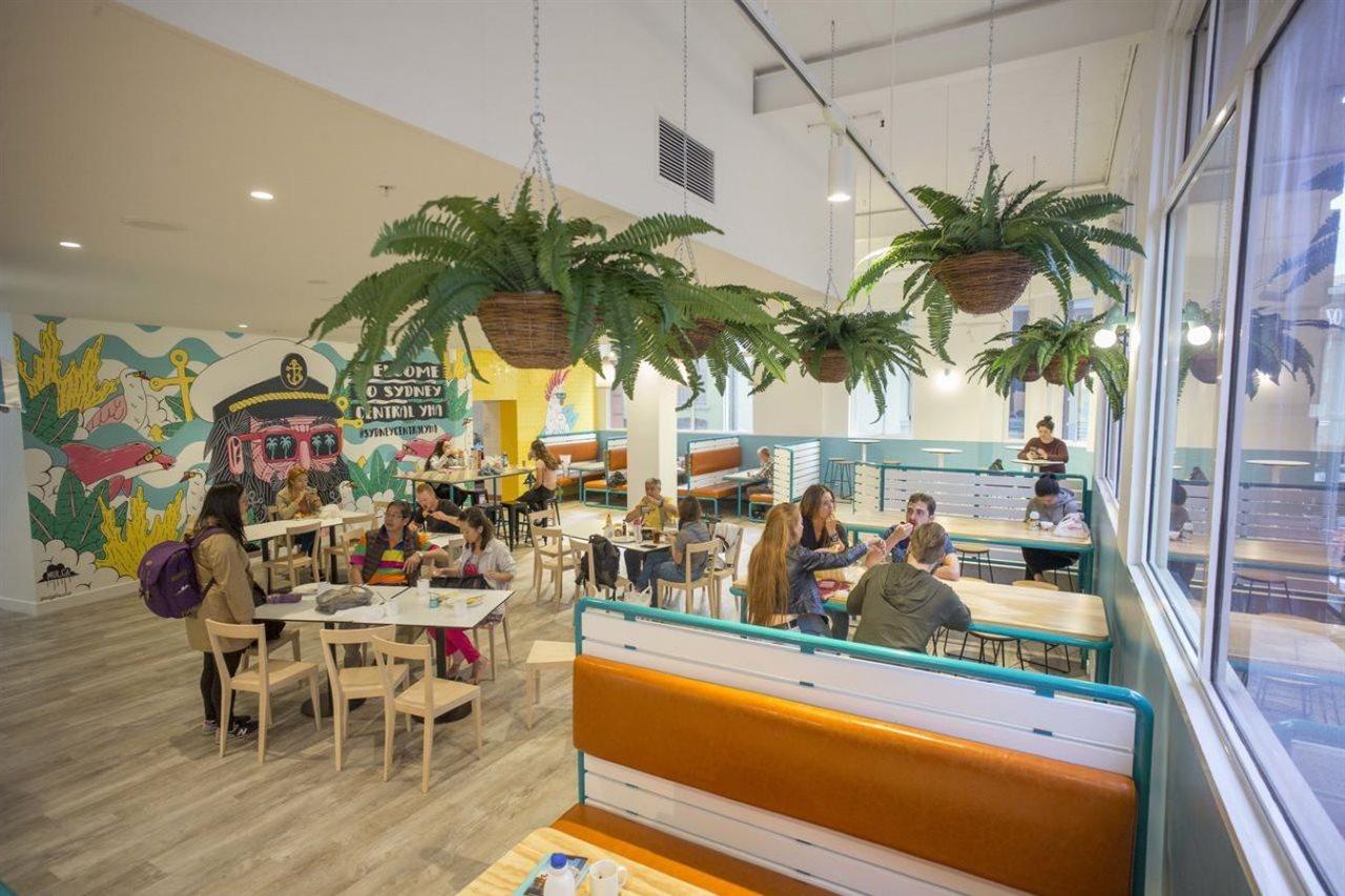 Yha Sydney Central Exterior photo The photo shows a vibrant and spacious café or restaurant interior. It features modern and casual seating arrangements, including tables and a booth area with orange cushions. The space is decorated with hanging potted ferns and a colorful mural on o
